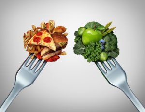 Diet struggle and decision concept and nutrition choices dilemma between healthy good fresh fruit and vegetables or greasy cholesterol rich fast food with two dinner forks competing to decide what to eat.